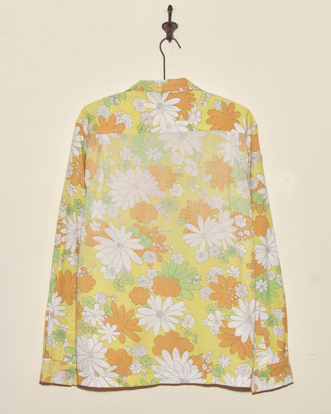 The Storytime Floral Shirt (XL/XXL), made from a vintage bedsheet, showcases a unique yellow, green, white, and orange floral pattern with long sleeves, and hangs gracefully on a hook.