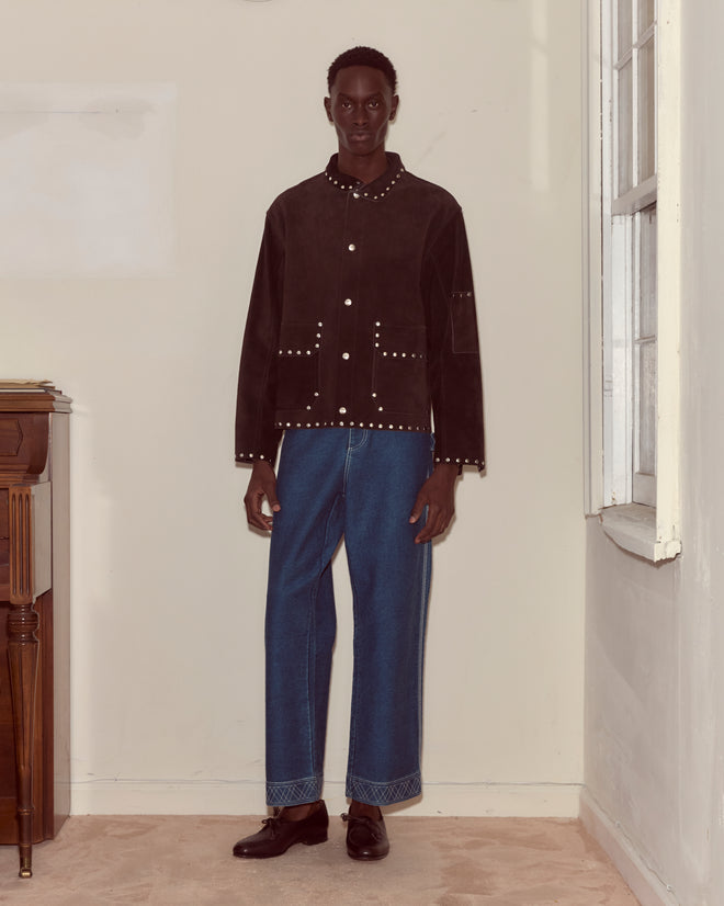 Indoors, a person stands by a window and piano wearing the Studded Welding Jacket - Brown with wide blue jeans.