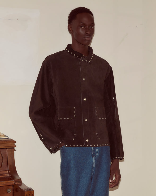 A person in a brown Studded Welding Jacket and blue jeans stands indoors against a plain wall.