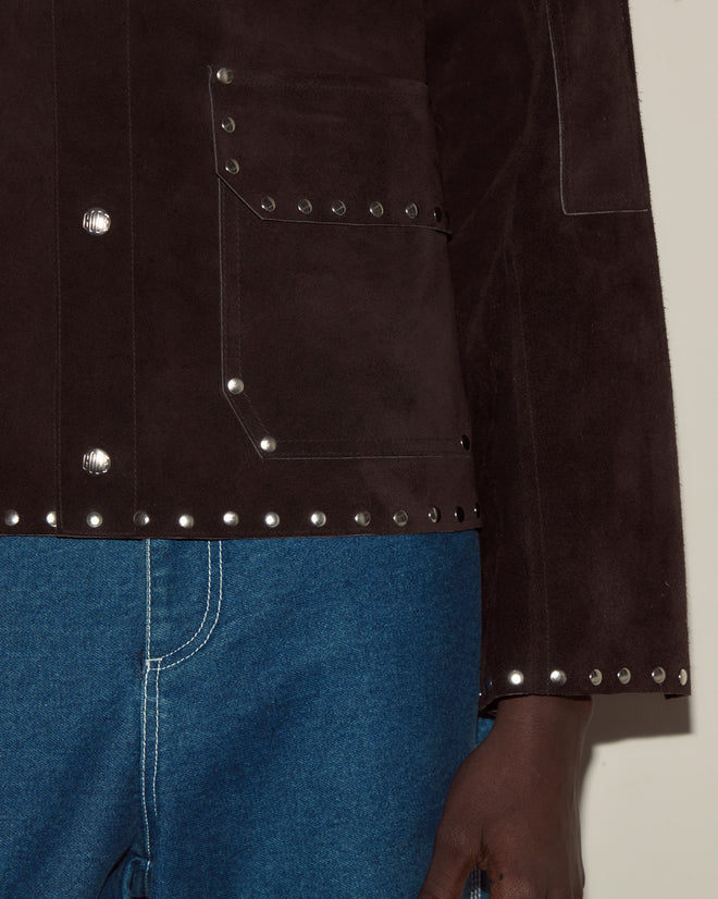 Someone is wearing the Studded Welding Jacket - Brown, a dark brown suede jacket with metal studs, paired with blue jeans and their hand by their side.