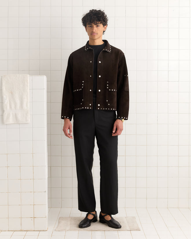 A person stands in a tiled room wearing the Studded Welding Jacket - Brown, paired with black pants and black shoes.
