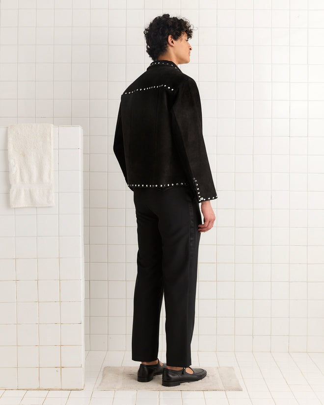 A person wearing the Studded Welding Jacket - Black stands on a mat in a white-tiled room, facing away from the camera.