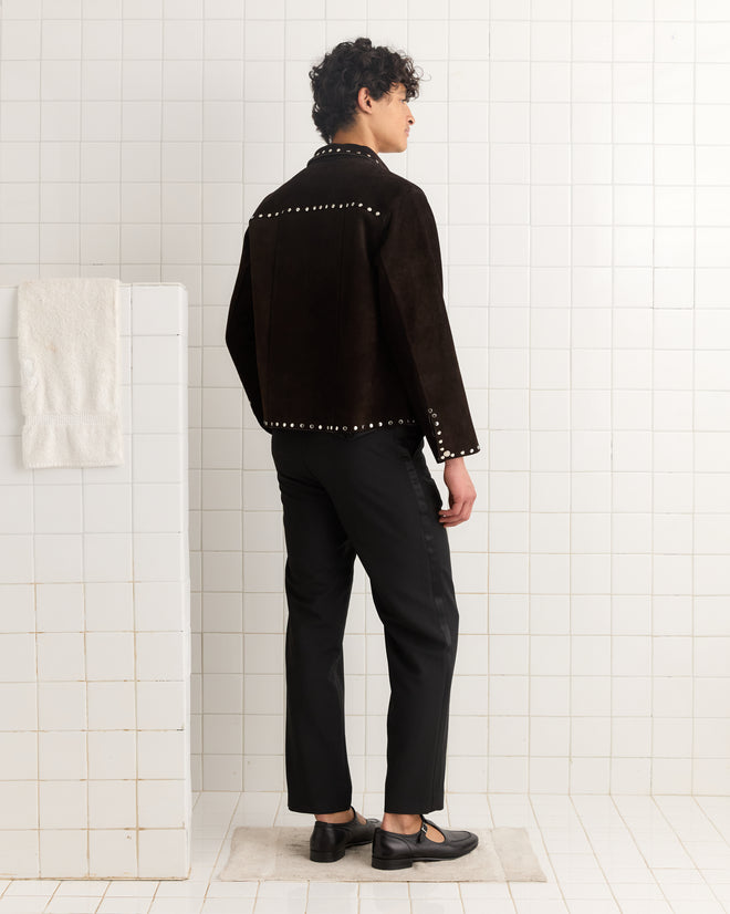 A person stands on a mat in a tiled room, facing away, wearing the Studded Welding Jacket - Brown with black pants and black shoes.