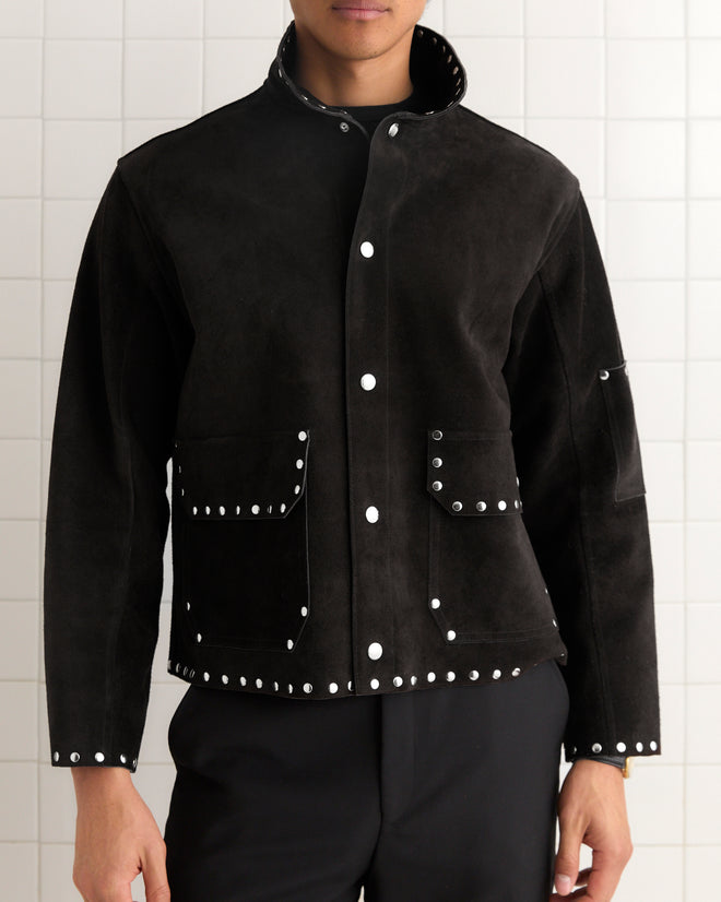 A person stands against a tiled wall wearing the Studded Welding Jacket - Black, featuring silver studs and front pockets, inspired by a leather specialist’s tailored style.