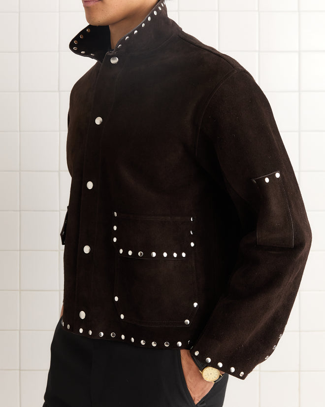 A man stands against a tiled wall wearing the Studded Welding Jacket - Brown, a dark suede jacket with silver studs inspired by metal shop welding accents.