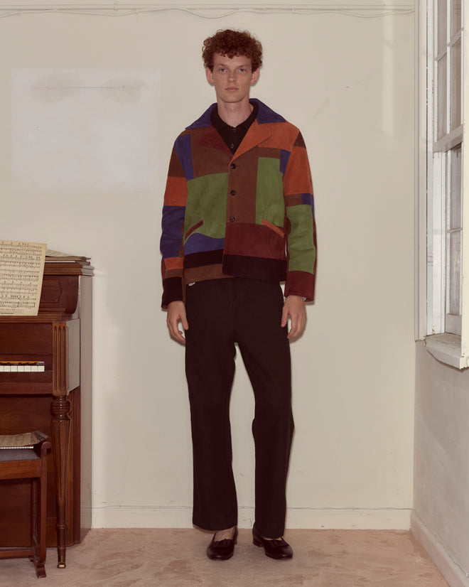 A man stands indoors beside a piano with sheet music, wearing black pants and the stylish Suede Collage Jacket, showcasing fashionable men's outerwear.
