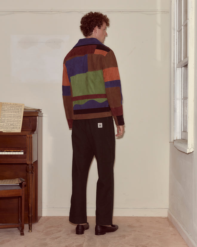 A person stands indoors near a piano, wearing black pants and the vibrant Suede Collage Jacket.