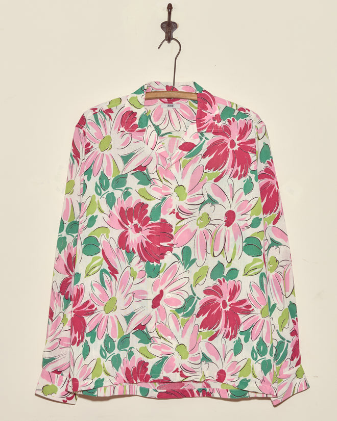 The Sukhothai Flower Shirt is a long-sleeve vintage top with a bold pink, white, and green floral print, displayed on a wall hook.
