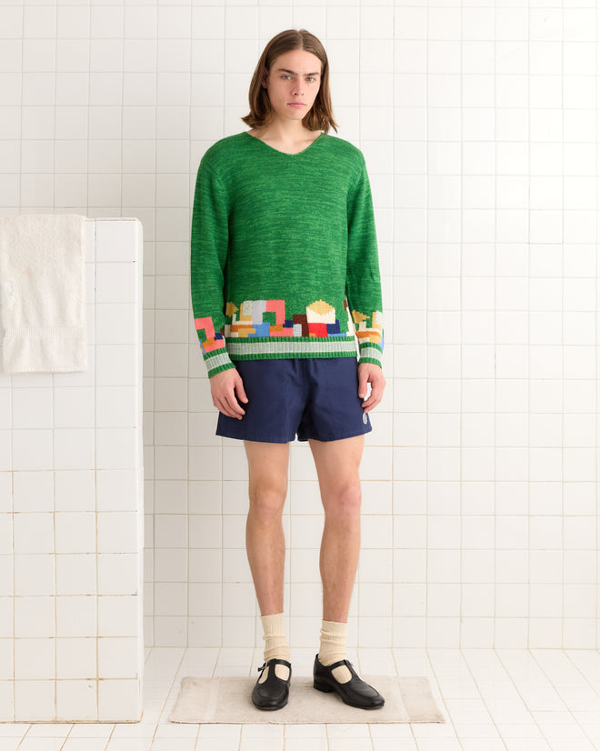 In a white-tiled room, a person stands wearing a Swatch Sweater in green with blue shorts, beige socks, and black shoes.