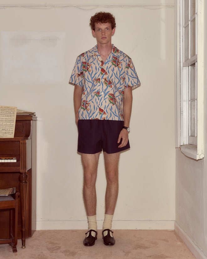 A man wearing the Swimmers Short Sleeve Shirt with a blue seaweed pattern stands indoors by a piano, looking at the camera.