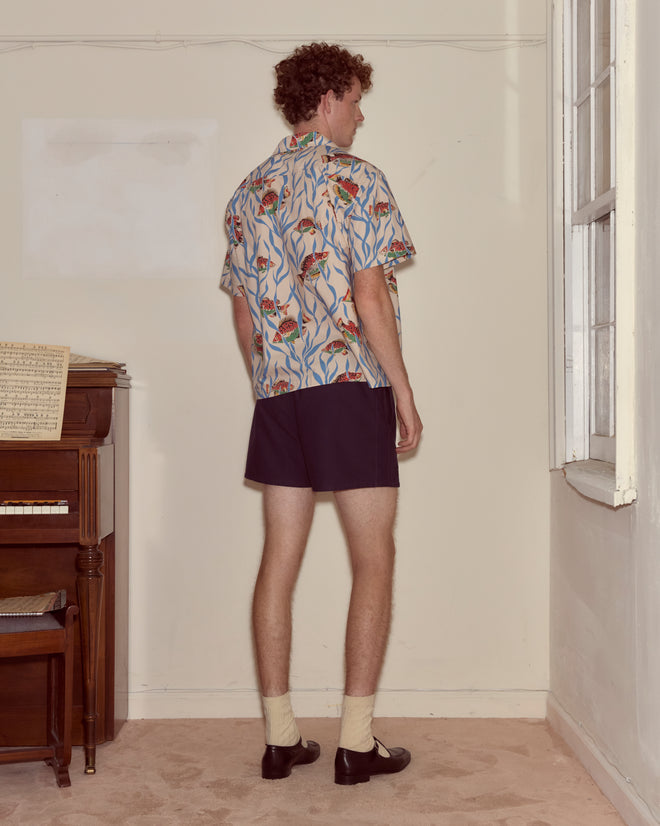 A man in a Swimmers Short Sleeve Shirt and shorts stands facing a wall near a piano and window in a vintage room.