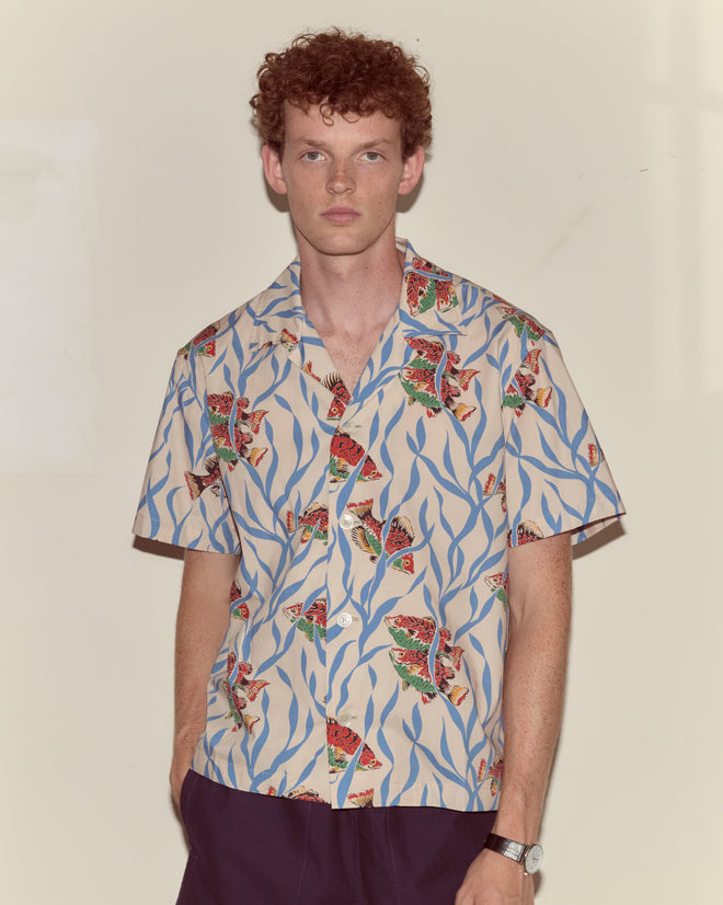 A man with curly hair wears the Swimmers Short Sleeve Shirt with a blue seaweed pattern and dark pants, standing against a plain background.