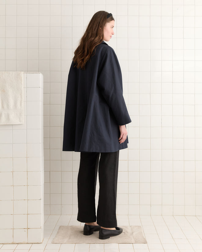 A person in a sleek Waxed Herringbone Peacoat and black pants stands in a white tiled room, facing sideways.