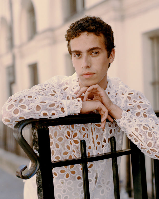 A young person in the Primrose Lace Long Sleeve Shirt, made from 100% viscose, leans on a black metal fence and gazes thoughtfully at the camera.