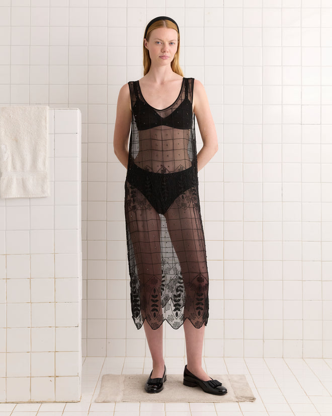 A woman wears the Beaded Grid Vine Dress - Black, a sheer black silk crepe dress with a geometric vine pattern, and black flats while standing in a white tiled room with a towel hanging on the wall.