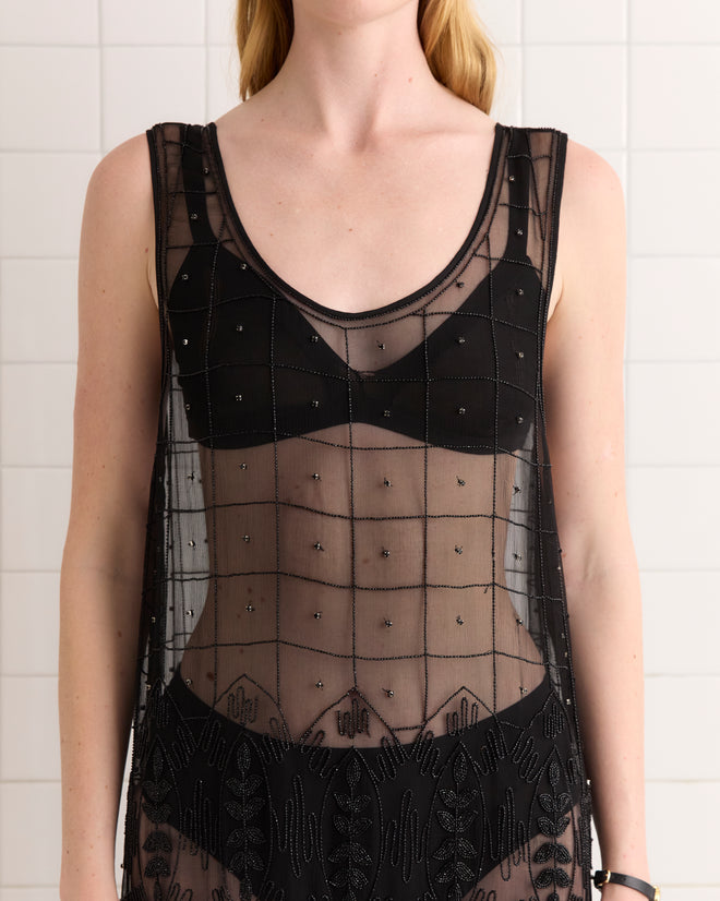 A woman wears the Beaded Grid Vine Dress - Black, a sheer 1920s-inspired dress with a geometric vine pattern over a black bra, standing before a tiled wall.