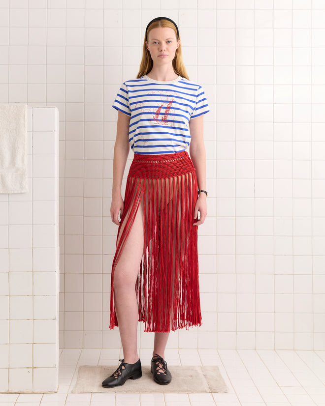 Woman standing in a tiled room wearing a striped t-shirt, black shoes, and the Crochet Fringe Skirt - Red.