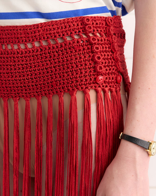 A person wearing a Crochet Fringe Skirt - Red, a striped shirt, and a black wristwatch.