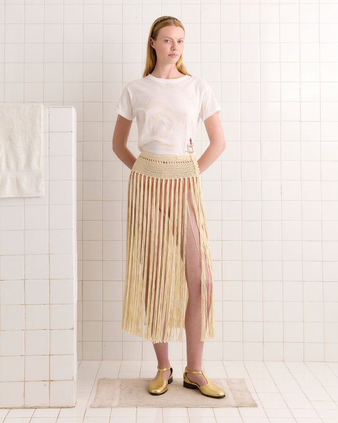 Woman in a white t-shirt and Cream Crochet Fringe Skirt stands on a mat in a tiled bathroom, wearing gold shoes.