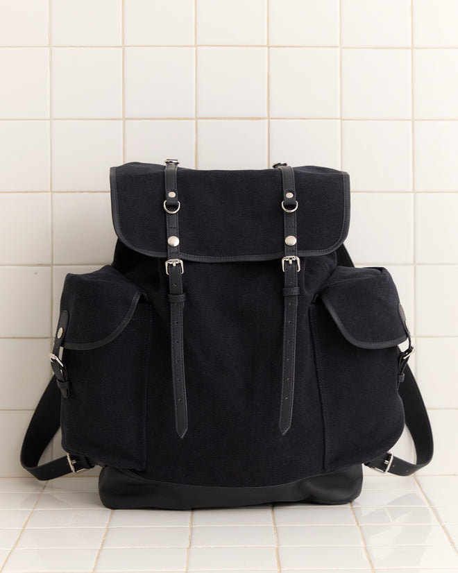 The Canvas Rucksack - Black, crafted from heavyweight canvas with front buckles and side pockets, is displayed against a white tiled background.