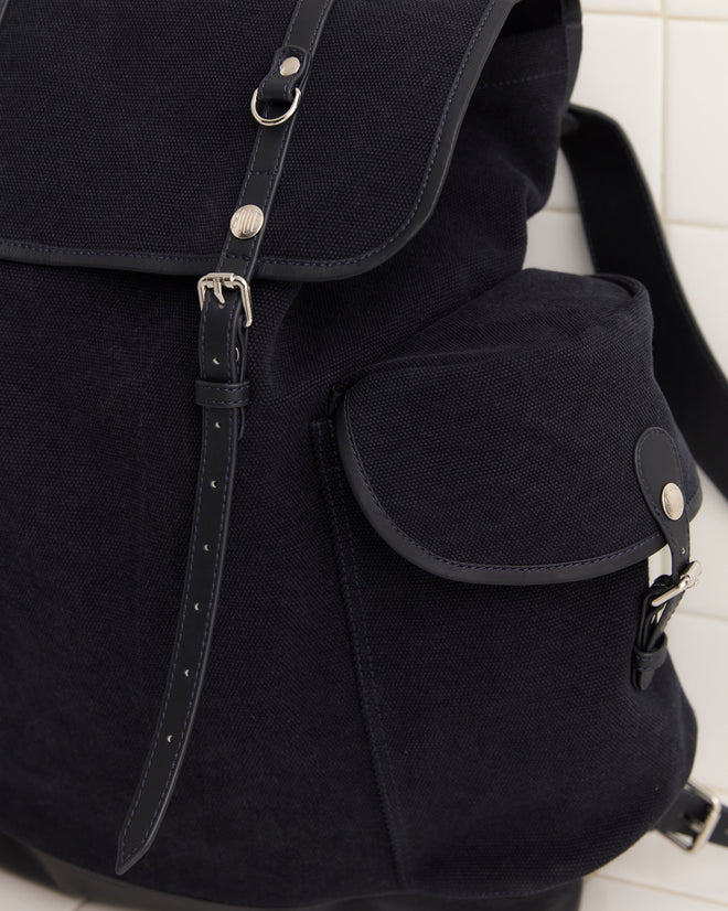 Close-up of the Canvas Rucksack - Black, crafted from heavyweight canvas with leather accents and a small side pocket, set against a tiled wall.