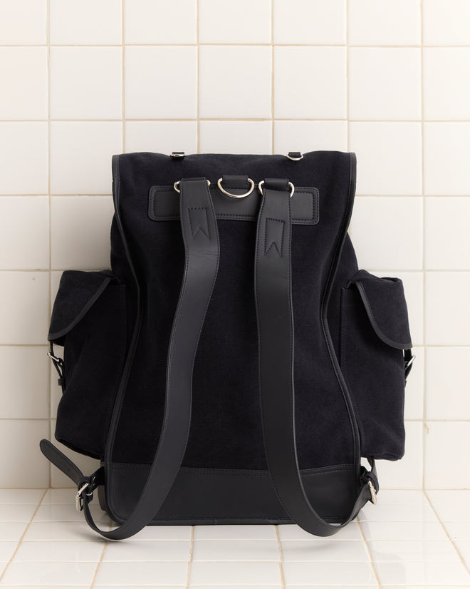 The Canvas Rucksack - Black, featuring leather accents, two front straps, and side pockets, is displayed against a tiled wall.