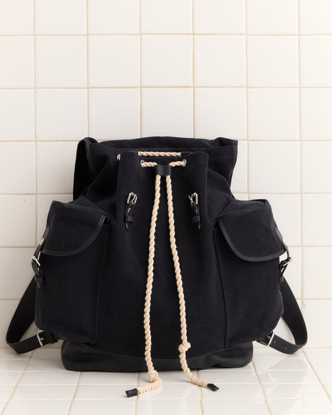 The Canvas Rucksack - Black features heavyweight canvas, leather accents, side pockets, and a white rope drawstring, shown against a tiled wall.