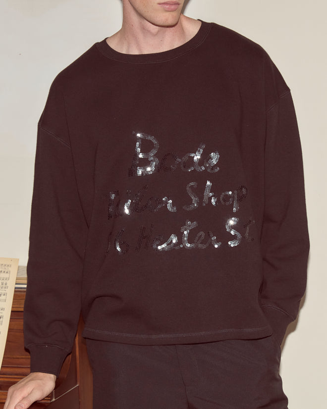Man wearing the Tailor Shop Crewneck - Black, a dark sweatshirt with shiny, partially readable text on the front, standing indoors.
