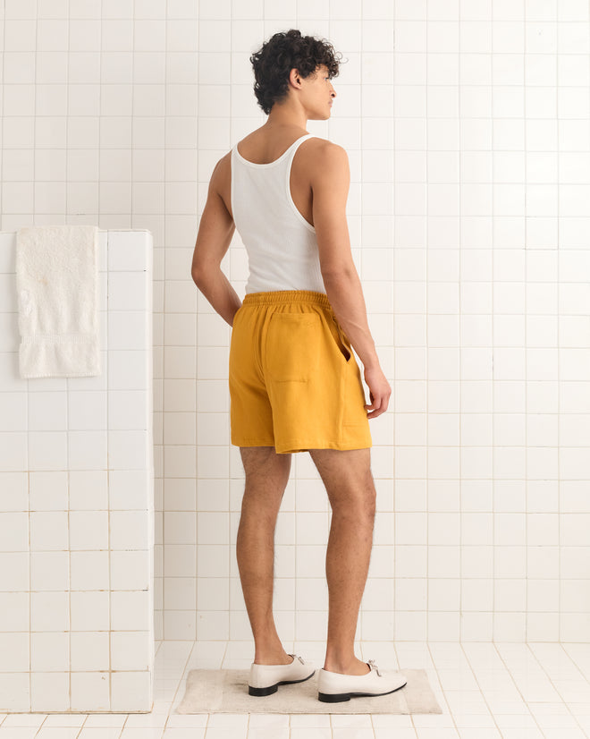 Man in a white tank top and Tennis Ball Patch Shorts stands on a mat in a white tiled bathroom, facing away.