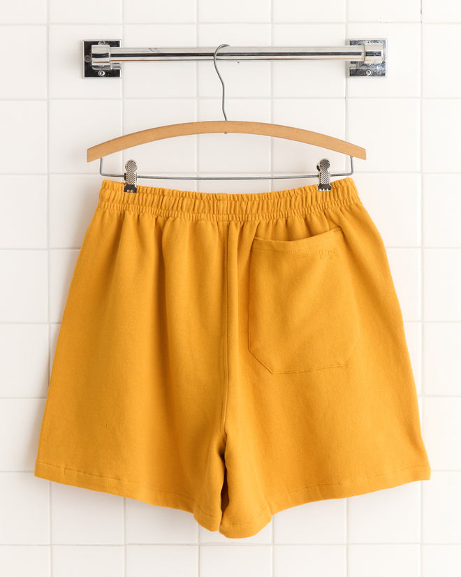 Tennis Ball Patch Shorts in yellow, made from 100% cotton with an elastic waistband and back pocket, displayed on a wooden hanger against a white tiled wall.