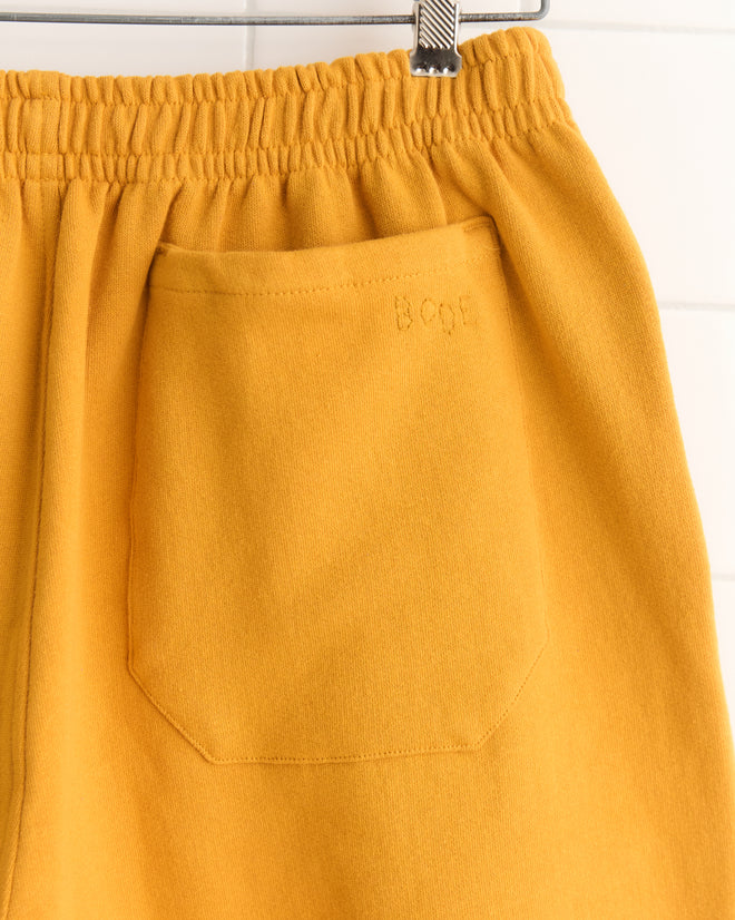Close-up of Tennis Ball Patch Shorts in mustard yellow 100% cotton, featuring an elastic waistband and a back pocket, hanging on a hanger.