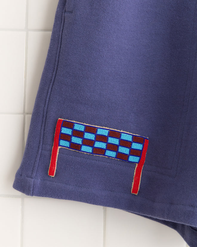 Close-up of Tennis Player Shorts in blue, featuring a red and blue checkerboard patch near the hem, reminiscent of Canadian rugby shorts, displayed on a tiled background.