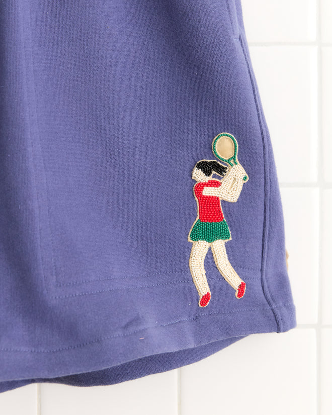 Tennis Player Shorts: Blue fabric sweatshorts featuring an embroidered patch of a tennis player in a red top, green skirt, and holding a racket.