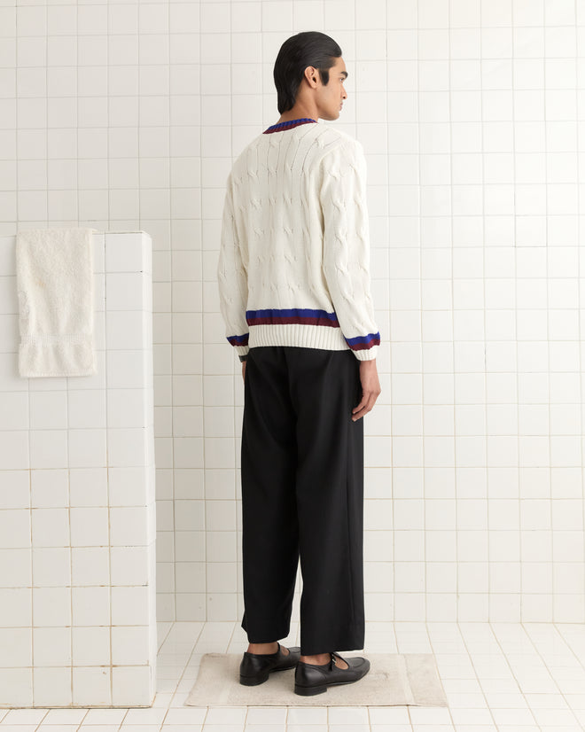 Dressed in a Tennis Sweater with navy and burgundy stripes, a person stands in a tiled room facing away, their black pants completing the look.