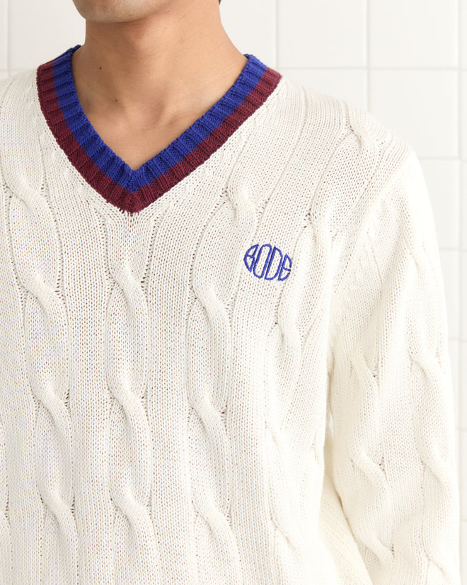 Someone wears the Tennis Sweater, a cream cable-knit pullover featuring blue and maroon collar accents and an embroidered Bode logo.