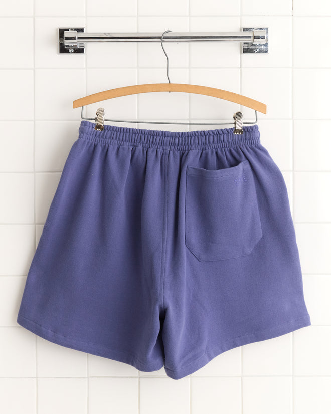 Tennis Player Shorts in purple with an elastic waistband and back pocket, displayed on a wooden hanger against a tiled wall.