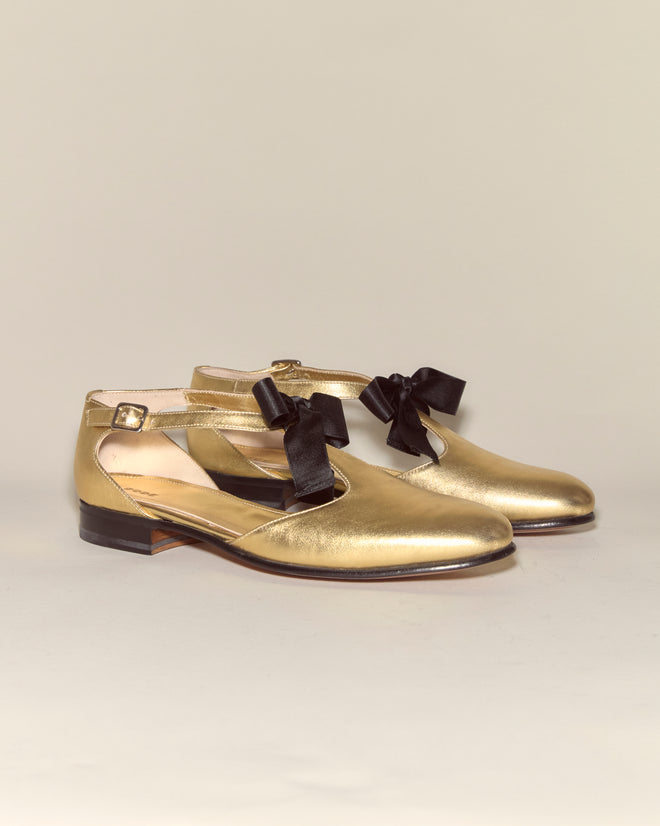 Theater Shoes - Gold feature gold metallic leather with ankle straps and black ribbon bows, reminiscent of classic midcentury dancing shoes, displayed on a neutral background.