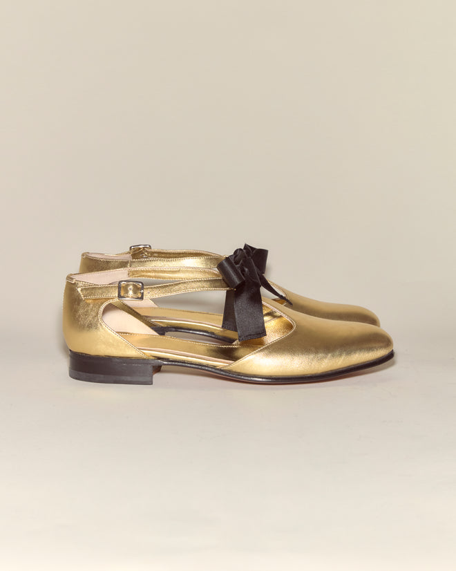 Theater Shoes - Gold: Metallic leather shoes with black bows, ankle straps, and low heels recall classic midcentury dance styles, shown side by side on a neutral background.