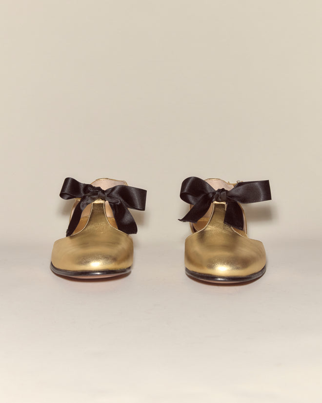 Theater Shoes - Gold: A pair of gold metallic leather shoes with black ribbon bows, inspired by classic midcentury dancing footwear, displayed against a plain light background.