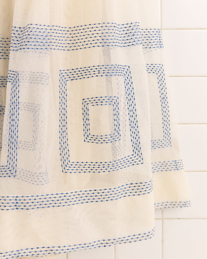 The Tile Stitch Dress, a white dress featuring geometric blue square embroidery, is displayed against a white tiled wall.
