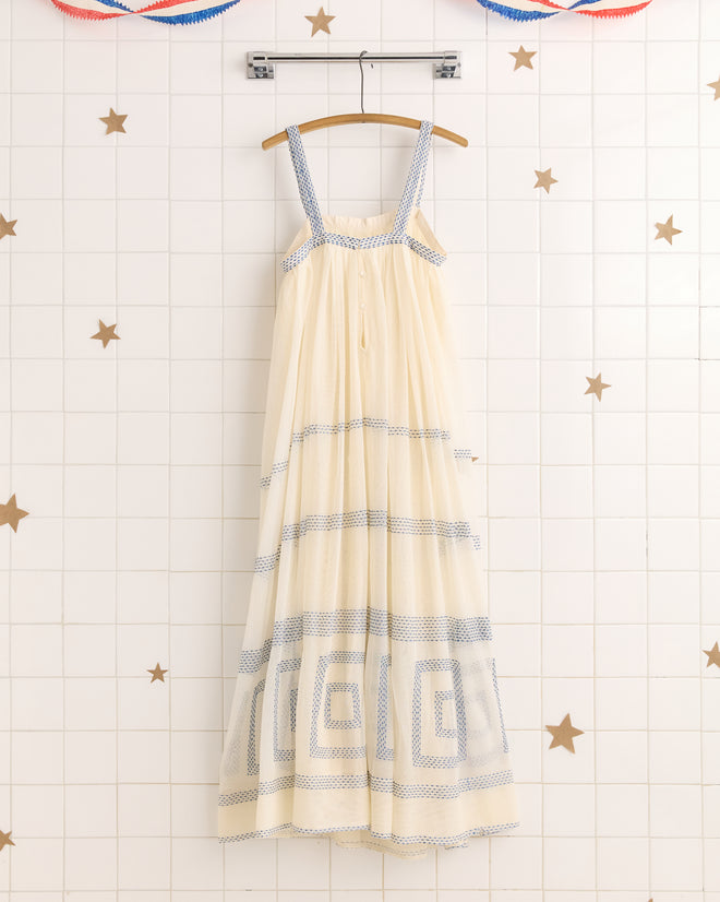 The Tile Stitch Dress is a cream sleeveless maxi dress inspired by the 1930s, featuring geometric blue embroidery. Shown on a wooden hanger, it stands out against a star-tiled wall.