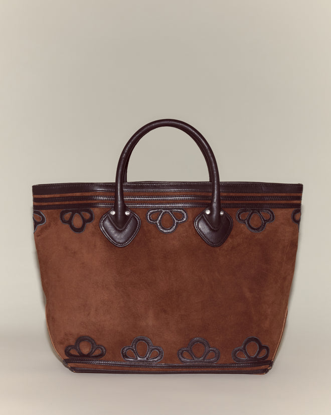 The Trefoil Tote - Brown is a boxy brown suede tote with dark brown leather handles and decorative leather appliqué accents on a neutral background.