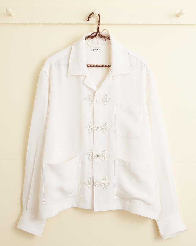 The Trillium Long Sleeve Shirt - White is a 1940s-inspired silk crepe shirt with trompe l'oeil knot buttons and three front pockets, shown hanging on a hook against a cream wall.