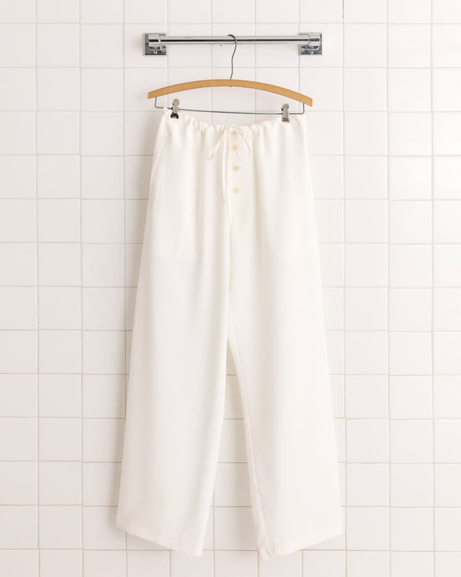 Cream Trillium Pajama Pants with a wide-leg silk design, elastic waistband, adjustable drawstring, and buttons hang on a wooden hanger against a tiled wall for a subtle 1950s-inspired look.
