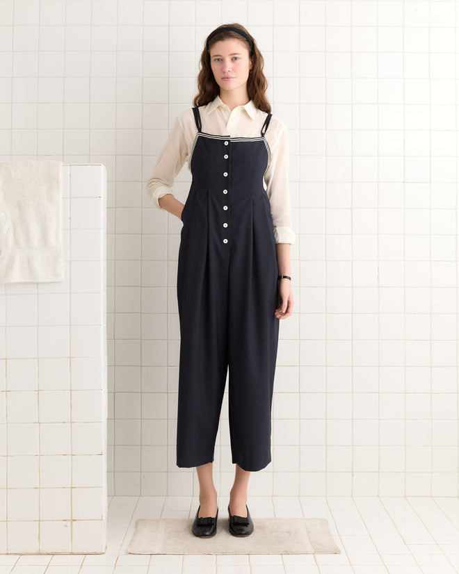A person in the Trimmed Gardner Jumpsuit - Navy and a white shirt with antique pearl buttons stands on a tiled floor against a white tiled wall.