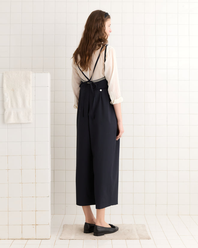 A woman in a white blouse and the Trimmed Gardner Jumpsuit - Navy, featuring antique pearl buttons, stands in a white-tiled room.