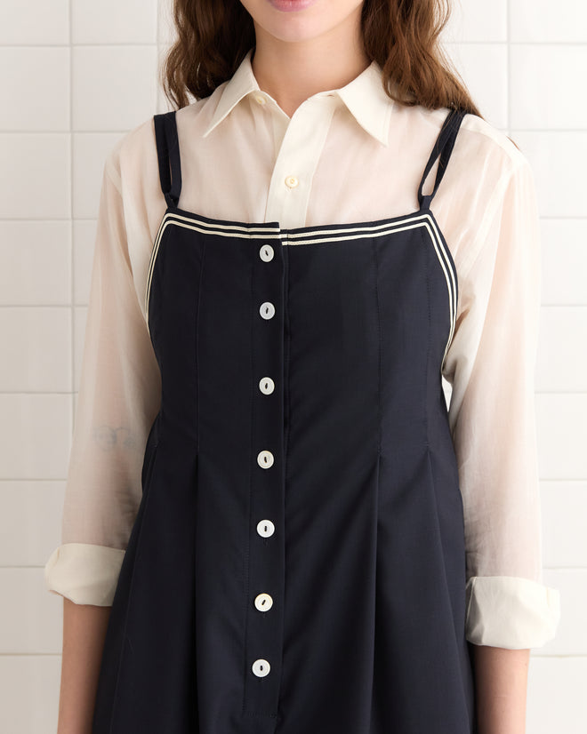 A person in a navy Trimmed Gardner Jumpsuit, adorned with antique pearl buttons over a white blouse, stands against a tiled wall, exuding timeless elegance.