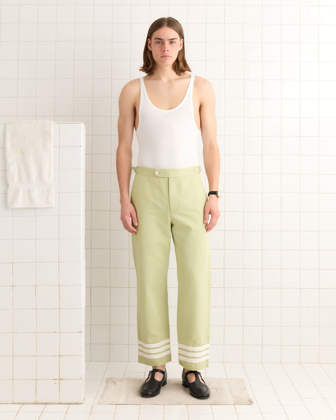 In a white tiled room, a person wearing a white tank top pairs their mid-rise trousers with Tri-Stripe Trousers in Green/Cream, showcasing subtle woven appliqué stripes for added charm.