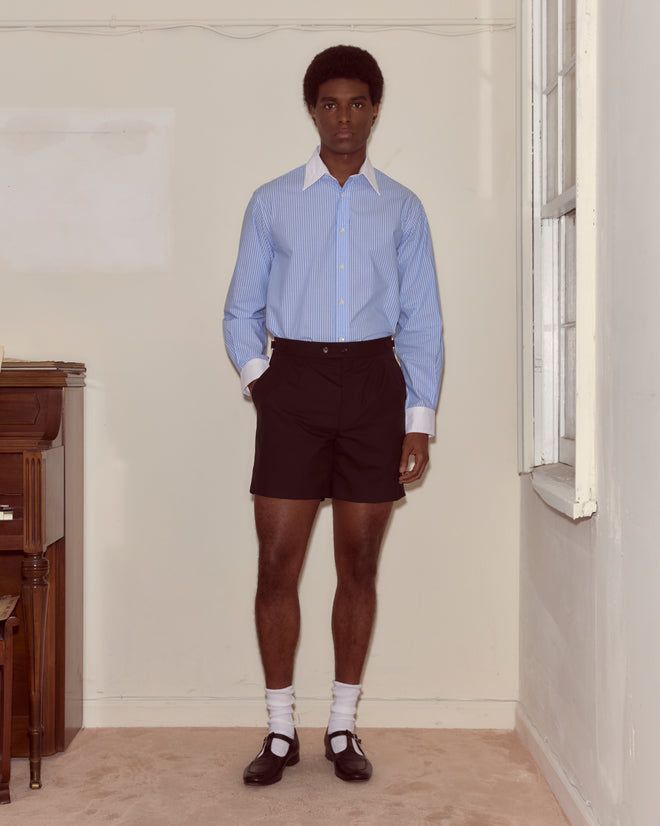 A person stands indoors wearing a blue striped shirt, Tropical Wool Double Button Shorts, white socks, and black shoes.