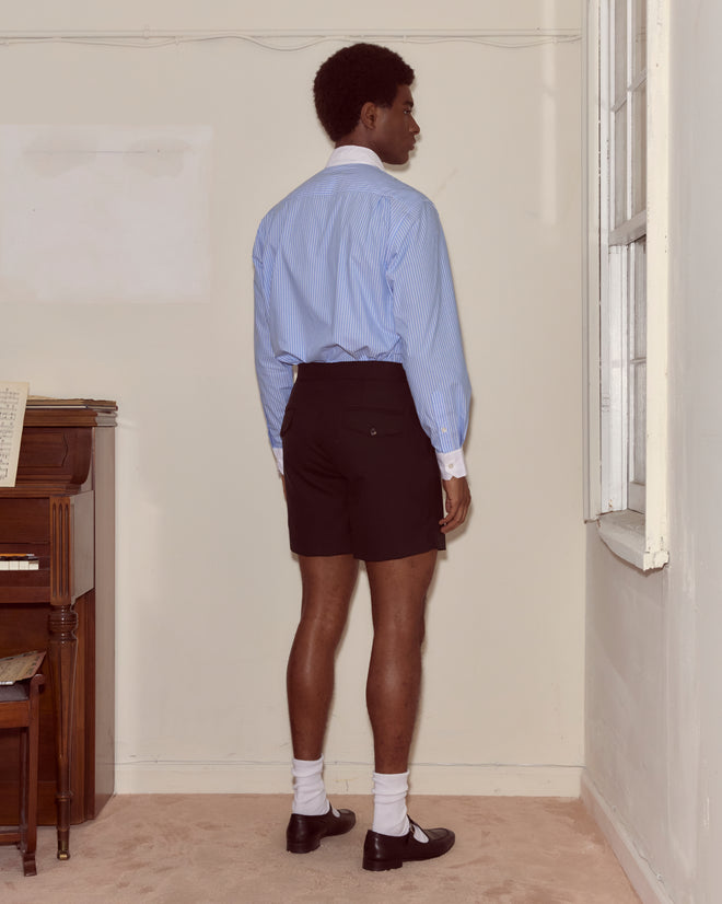 A man in a blue shirt and Tropical Wool Double Button Shorts stands facing a corner in a sparsely furnished room.